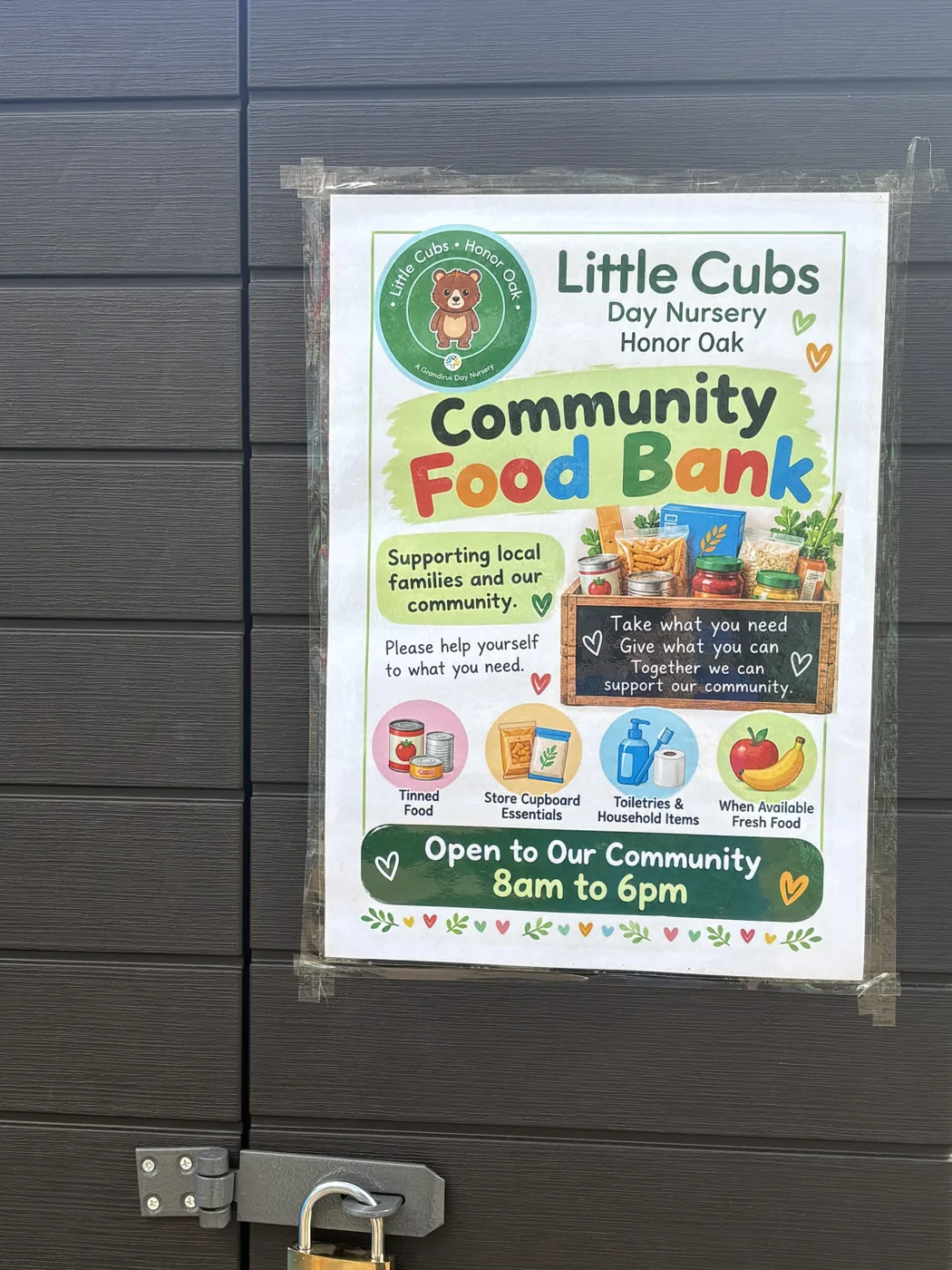 Poster on a dark grey cupboard door reading Little Cubs Day Nursery Honor Oak, Community Food Bank, supporting local families and our community, take what you need, give what you can, open to our community 8am to 6pm